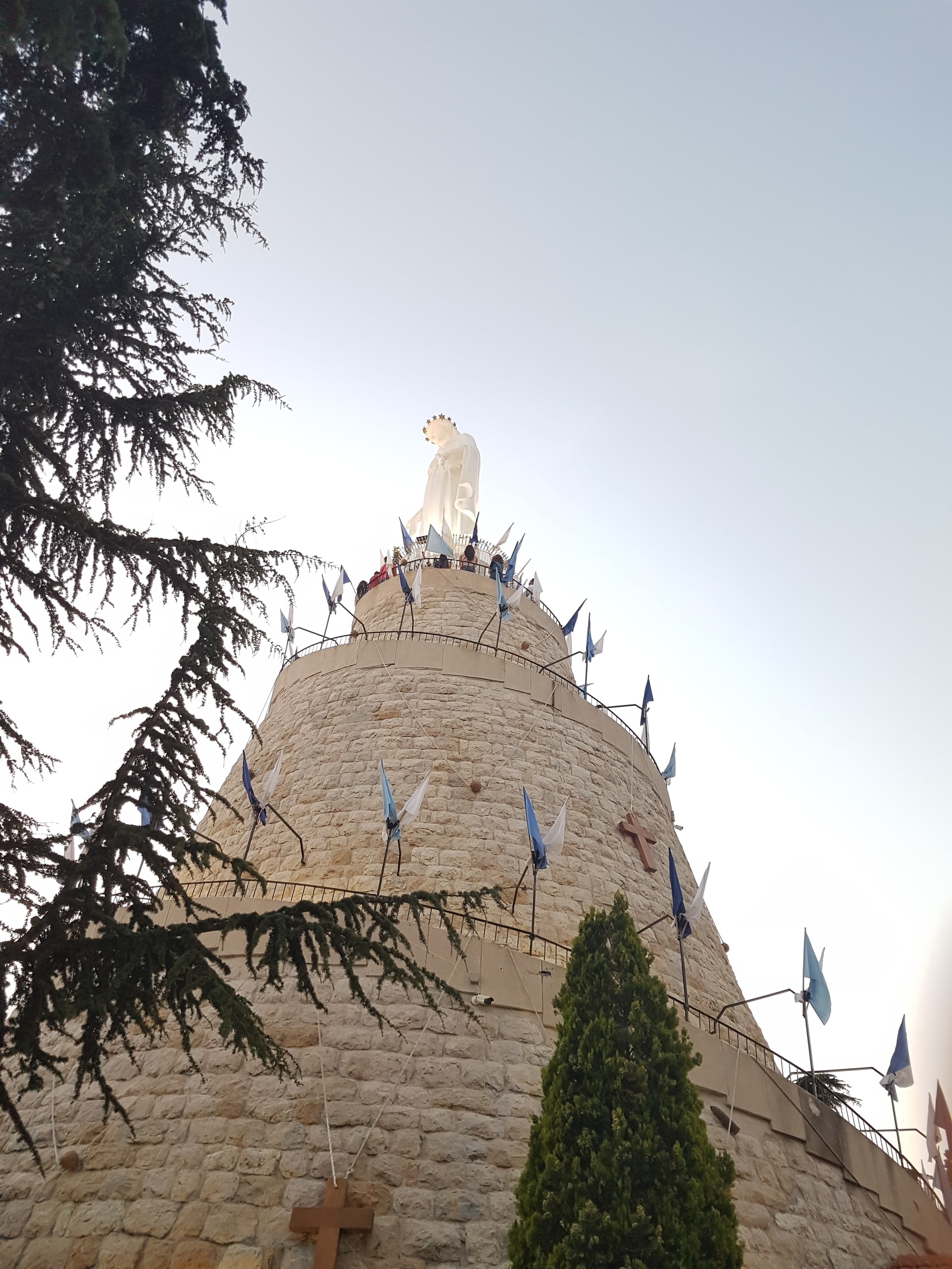 Stone tower hung with flags and crowned by a white statue, seen from below through cedar branches