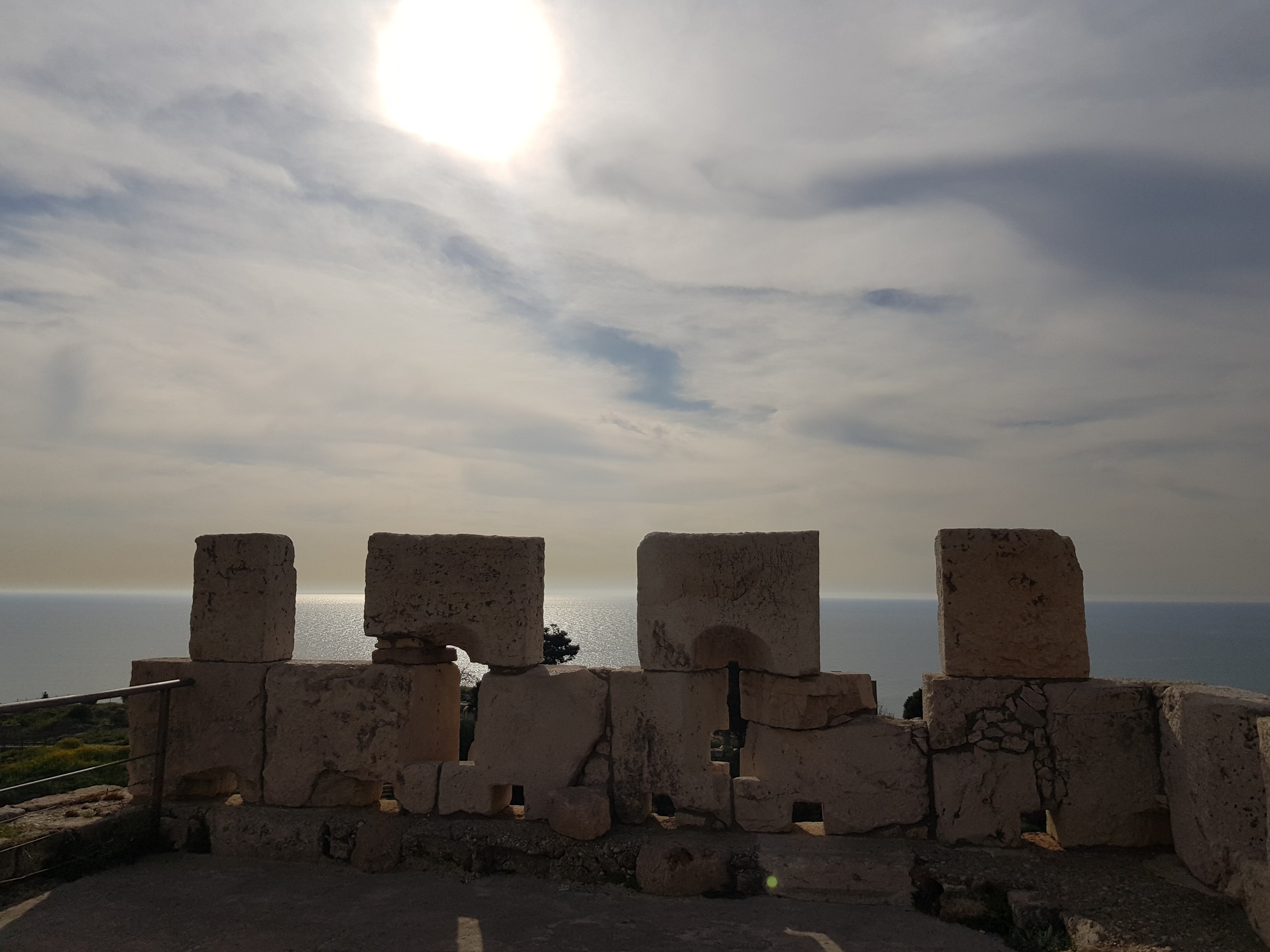 Ancient stone blocks silhouetted against a hazy sea, the sun behind cloud