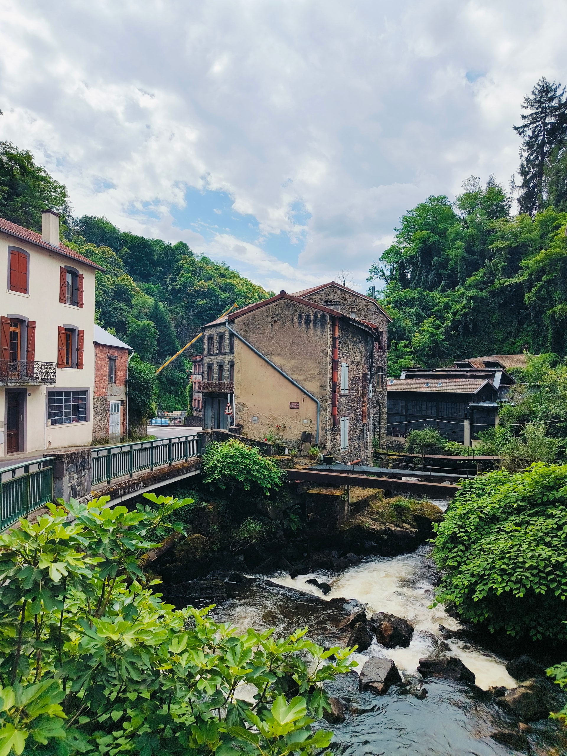 Stream tumbling beneath a footbridge between old houses and green trees