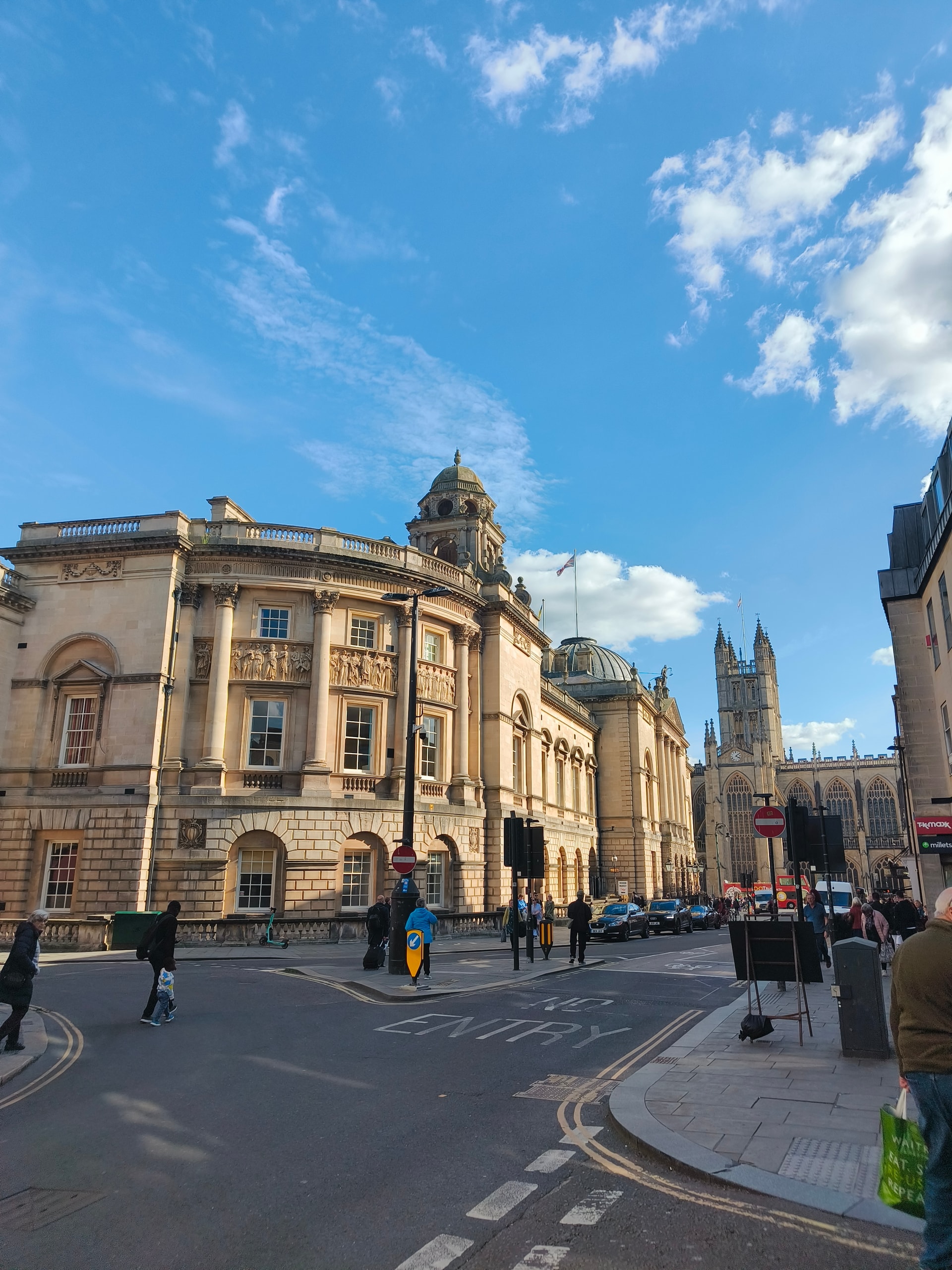 Honey-coloured stone buildings on a sunny street corner, an abbey tower in the distance