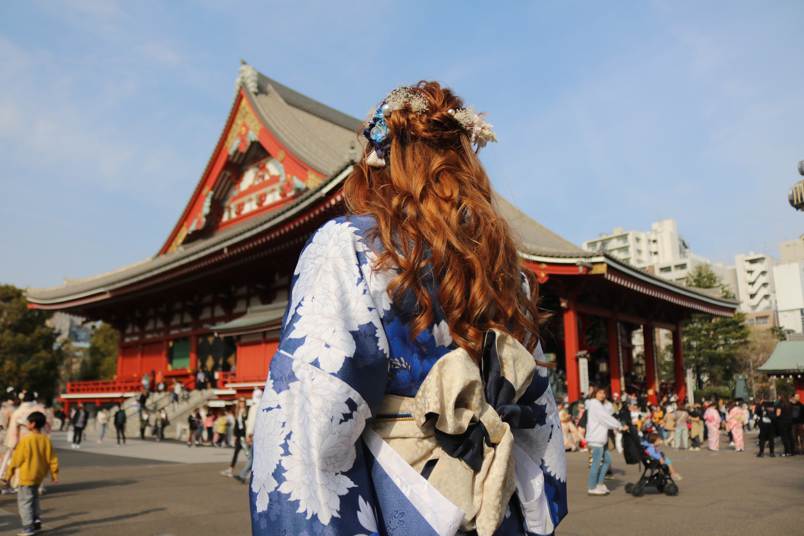 Woman in a blue floral kimono looking toward a red temple hall under a blue sky
