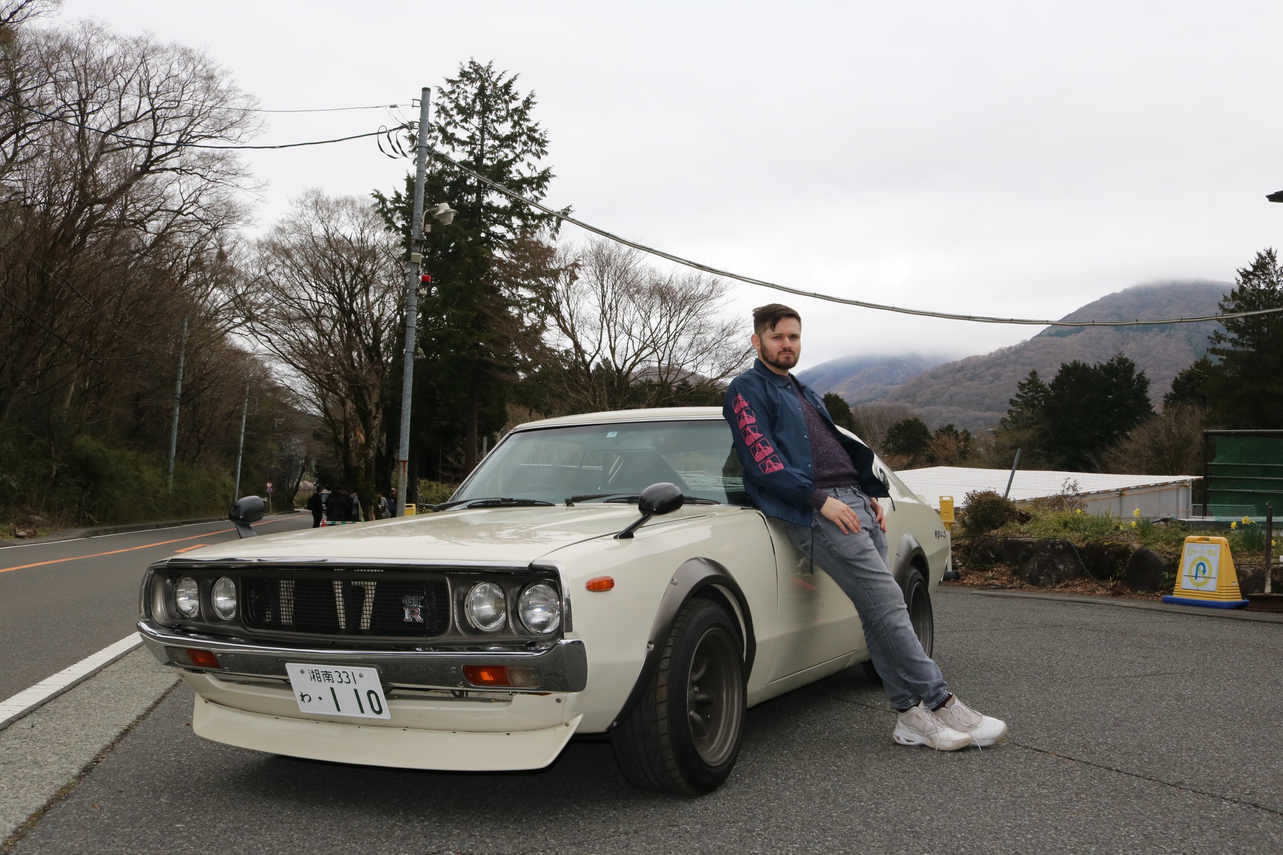 Man sitting on the front of a white vintage sports car at the roadside, hills behind