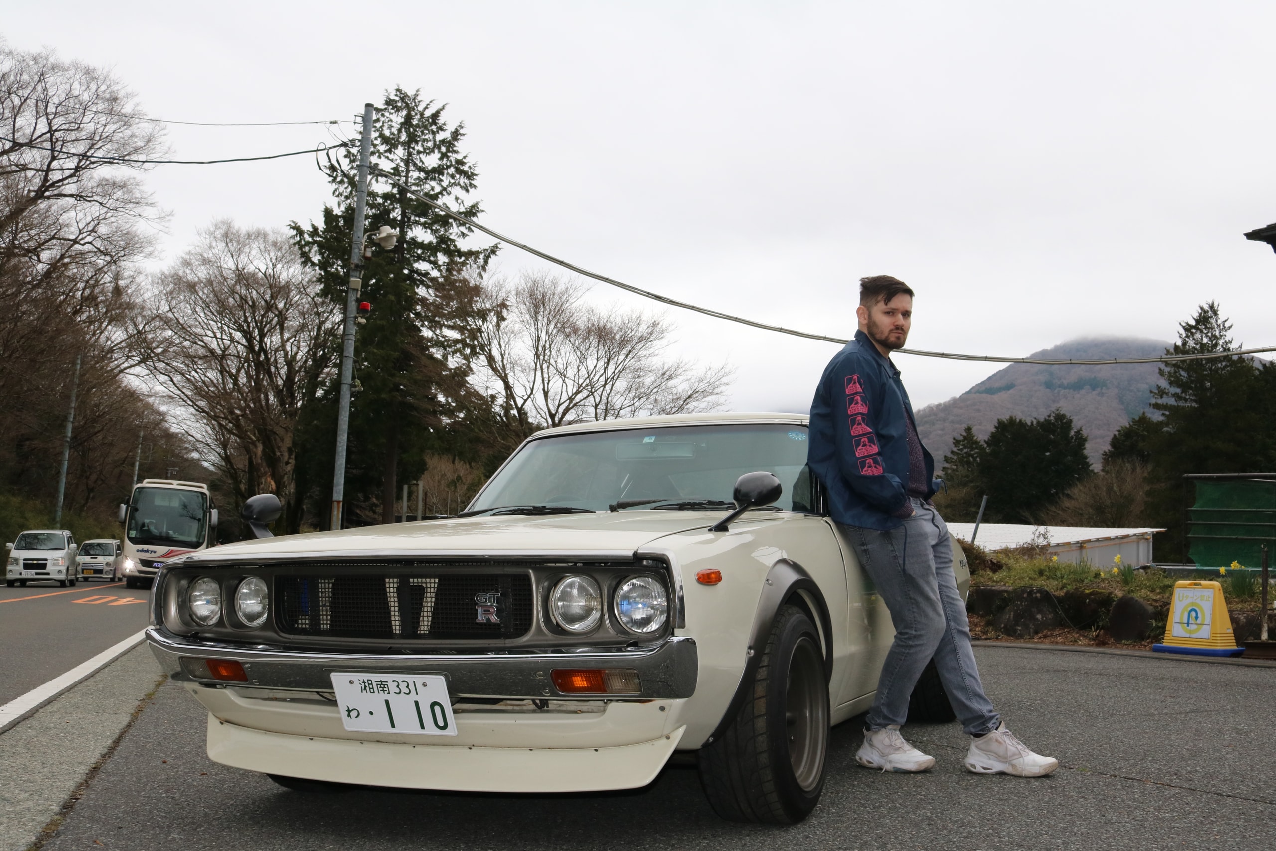 Man resting on the bonnet of a white vintage sports car beside a road, trees and hills behind