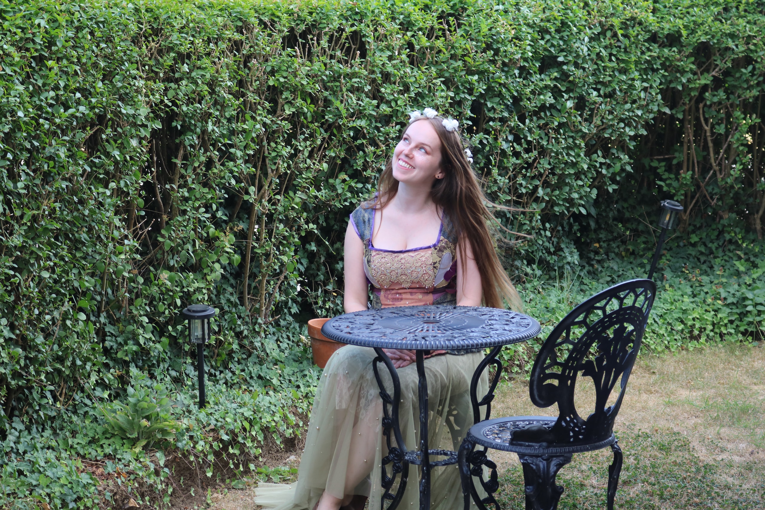 Woman in a medieval-style dress at an iron garden table in front of a hedge, looking up into the light