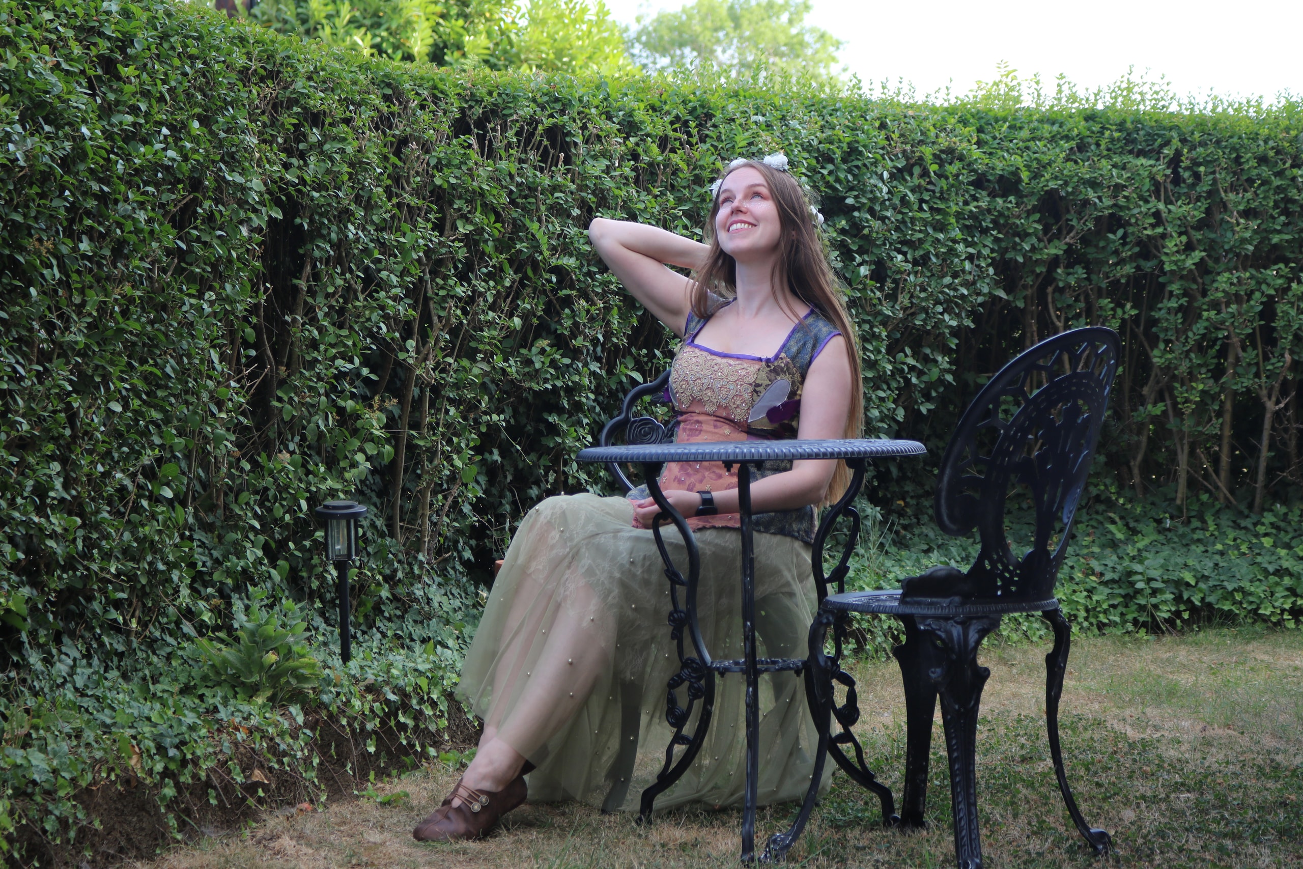 Woman in a medieval-style dress leaning back at a garden table with one arm raised, hedge behind her