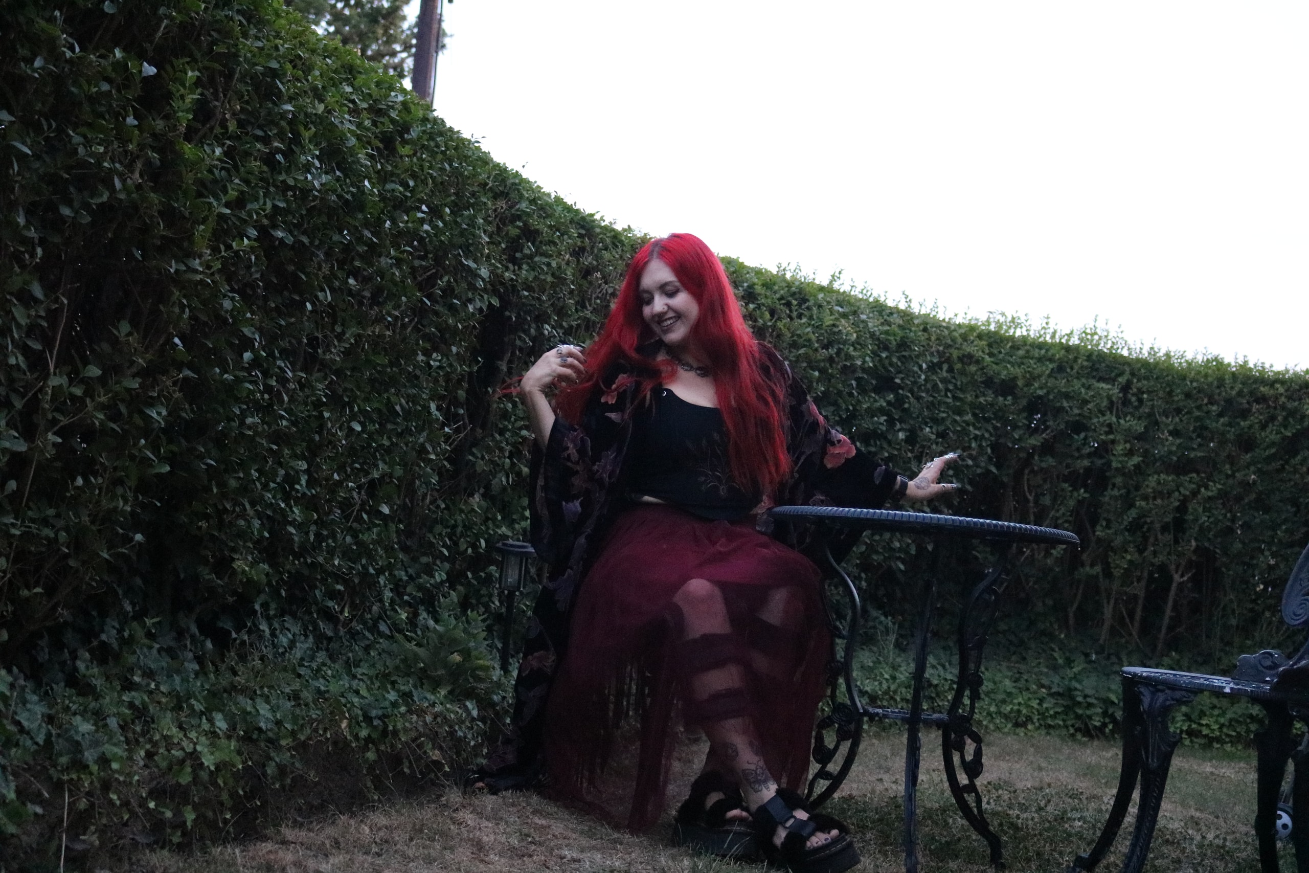 Woman with bright red hair in a dark red gown seated at an iron garden table beside a hedge