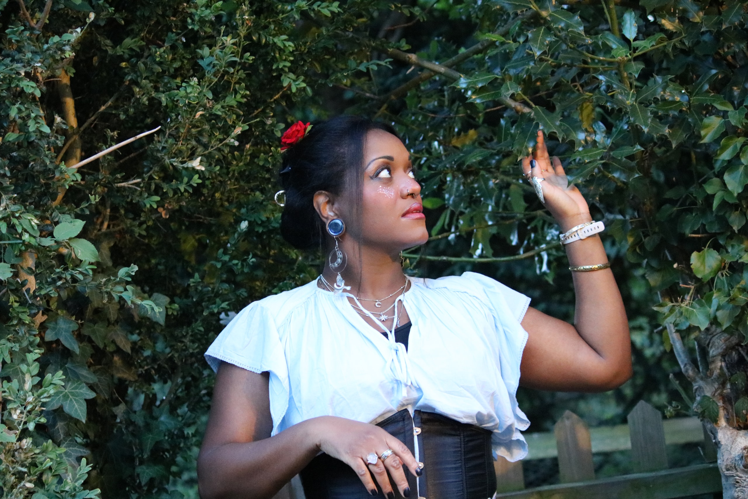 Woman in a white blouse with a red flower in her hair reaching toward overhanging leaves